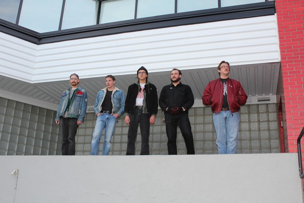 A colour photo of five-piece band standing in a line on a big concrete ledge outside of a building.