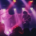 A colour photo shows three-piece band Oranje performing live bathed in red and purple lights. Due to the effect on the image, the bass player appears upside down. 