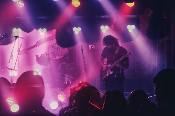 A colour photo shows three-piece band Oranje performing live bathed in red and purple lights. Due to the effect on the image, the bass player appears upside down. 