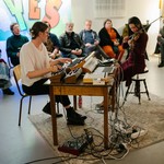 A colour photo shows duo Osmanthus performing while sitting down. One sits at a keyboard and the other plays the violin. A small audience is seen behind them watching the performance.