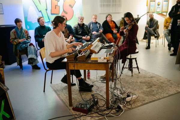 A colour photo shows duo Osmanthus performing while sitting down. One sits at a keyboard and the other plays the violin. A small audience is seen behind them watching the performance.