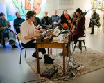 A colour photo shows duo Osmanthus performing while sitting down. One sits at a keyboard and the other plays the violin. A small audience is seen behind them watching the performance.