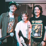 A colour photo shows three-piece rock band Pinball 1973 standing inside posing for a photo after a show. The middle member has their black and white guitar strapped on.