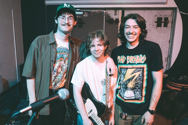 A colour photo shows three-piece rock band Pinball 1973 standing inside posing for a photo after a show. The middle member has their black and white guitar strapped on.