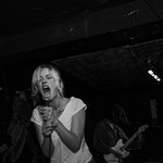 A black and white photo shows the singer of punk band PISS screaming into the microphone on stage at an indoor venue. They're wearing a white shirt and have blonde hair tied back. The guitar player is shown faintly behind them. 