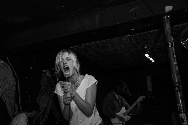 A black and white photo shows the singer of punk band PISS screaming into the microphone on stage at an indoor venue. They're wearing a white shirt and have blonde hair tied back. The guitar player is shown faintly behind them. 