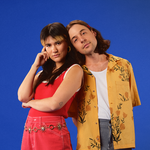 A bright colour photo shows the indie duo Pony Girl posing for a photo in front of a blue background. They're dressed in bright red and yellow clothes.