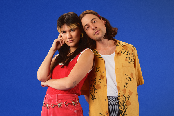 A bright colour photo shows the indie duo Pony Girl posing for a photo in front of a blue background. They're dressed in bright red and yellow clothes.