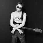 A black and white photo shows the artist Pressure Pin standing against a black bacground with their arms resting on their electric guitar. They're wearing a white t-shirt and circle sunglasses. 
