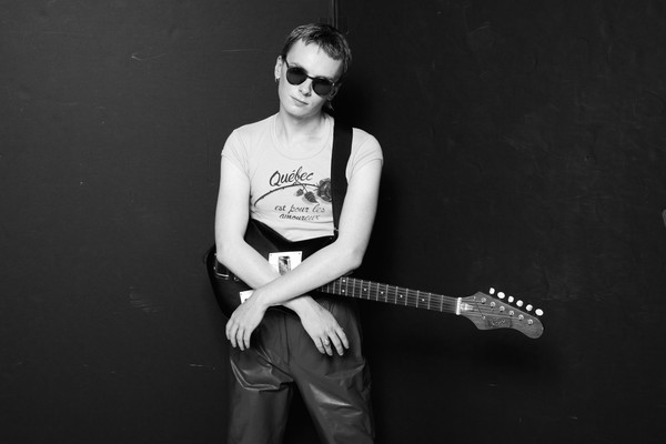 A black and white photo shows the artist Pressure Pin standing against a black bacground with their arms resting on their electric guitar. They're wearing a white t-shirt and circle sunglasses. 