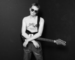 A black and white photo shows the artist Pressure Pin standing against a black bacground with their arms resting on their electric guitar. They're wearing a white t-shirt and circle sunglasses. 