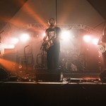 A colour photo shows four-piece rock band Puberty Well playing on a big lowlit stage under a kind of hazy orange light.