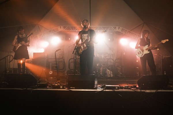 A colour photo shows four-piece rock band Puberty Well playing on a big lowlit stage under a kind of hazy orange light.
