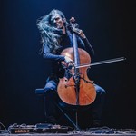 A colour photo shows cello Raphael Weinroth-Browne furiously playing a cello against a black backdrop. They are dressed all in black with long dark hair and various pedals and cords sit on the floor in front.