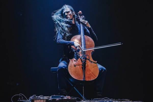 A colour photo shows cello Raphael Weinroth-Browne furiously playing a cello against a black backdrop. They are dressed all in black with long dark hair and various pedals and cords sit on the floor in front.