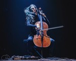 A colour photo shows cello Raphael Weinroth-Browne furiously playing a cello against a black backdrop. They are dressed all in black with long dark hair and various pedals and cords sit on the floor in front.