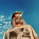 A colour photo taken from below shows artist Ryan Bourne with a bright blue sky behind them. They're wearing shimmering glasses, a tan shirt and a ball cap.