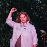 A colour photo shows artist Samantha Savage Smith standing outside in front of a bush. They're holding their right hand in the air above their head. 