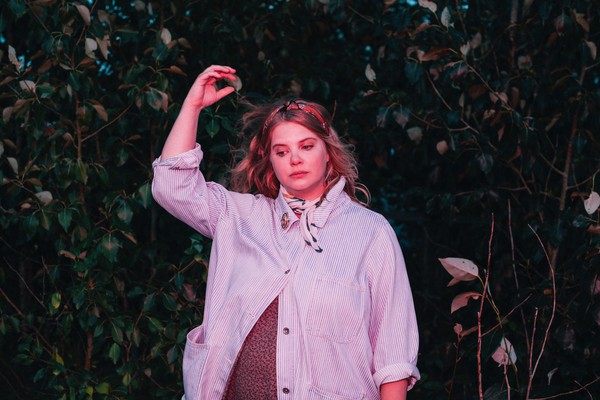 A colour photo shows artist Samantha Savage Smith standing outside in front of a bush. They're holding their right hand in the air above their head. 