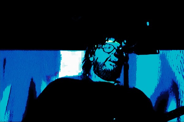 A blue and black photo shows artist SAMWOY singing into a microphone with a beard and sunglasses
