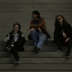 A dark lit colour photo shows three-piece group schoolyard sitting on a set of concrete stairs. The member on the left is seen smoking a cigarette, the member in the middle looks to their right, and the person on the right is looking straight at the camera.
