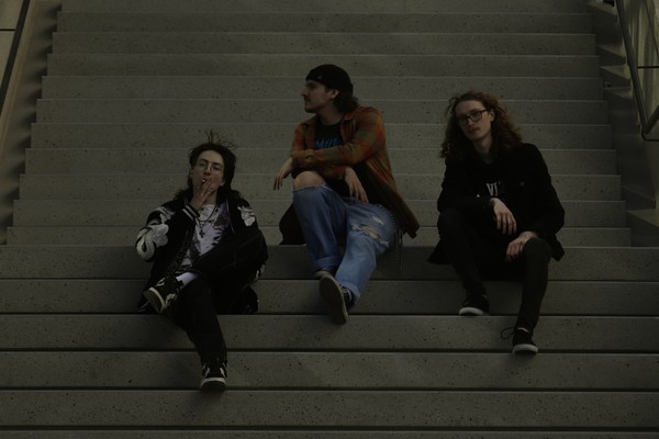 A dark lit colour photo shows three-piece group schoolyard sitting on a set of concrete stairs. The member on the left is seen smoking a cigarette, the member in the middle looks to their right, and the person on the right is looking straight at the camera.