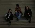 A dark lit colour photo shows three-piece group schoolyard sitting on a set of concrete stairs. The member on the left is seen smoking a cigarette, the member in the middle looks to their right, and the person on the right is looking straight at the camera.