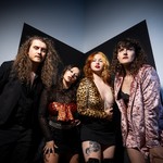 Four-piece band Shiv and the Carvers pose stand in a line and pose for a colour photo in the corner of a black and grey room