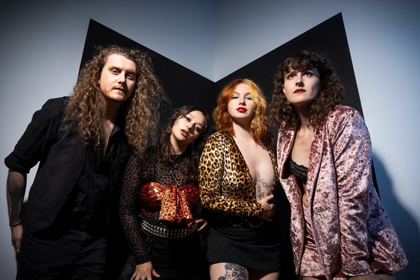 Four-piece band Shiv and the Carvers pose stand in a line and pose for a colour photo in the corner of a black and grey room