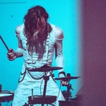 A grainy photo of artist SILKRØAD playing an electronc drum kit dressed in white clothes against a blue wall.