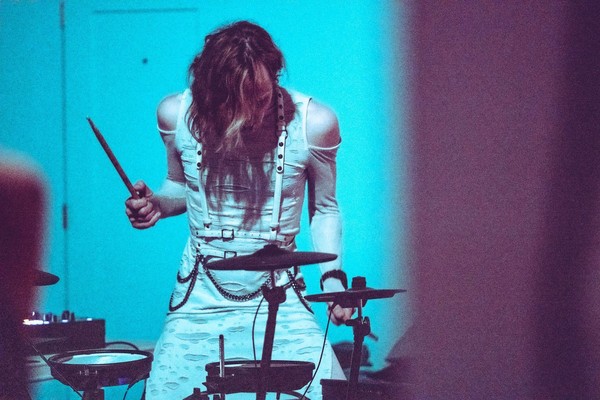 A grainy photo of artist SILKRØAD playing an electronc drum kit dressed in white clothes against a blue wall.