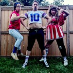 A colour photo of three-piece band SMOOCHER dressed as athletes and cheerleaders standing on the grass in a backyard against a wooden fence.