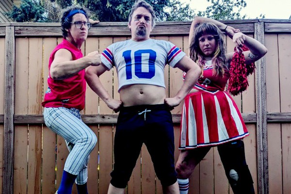 A colour photo of three-piece band SMOOCHER dressed as athletes and cheerleaders standing on the grass in a backyard against a wooden fence.