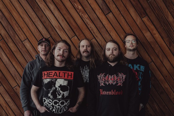 A colour photo of five-piece band Snakepit posing for a photo standing in front of an wall with angled wooden planks.