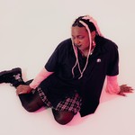 A colour photo shows artist So Sus sitting with their legs to the side in a white room. 