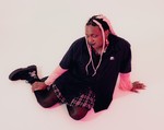 A colour photo shows artist So Sus sitting with their legs to the side in a white room. 