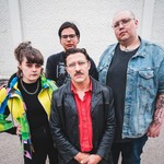 A colour photo of four-piece group Stucco shows them posing against a white brick wall.