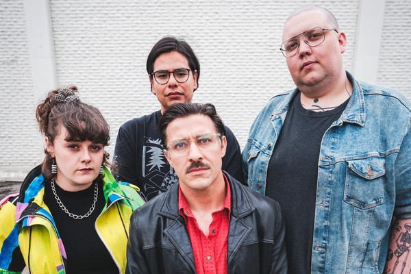 A colour photo of four-piece group Stucco shows them posing against a white brick wall.