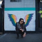 A colour photo shows the artist Sunflower Dreams crouching in front of a wall with wings painted on it, so it looks like they have wings. 