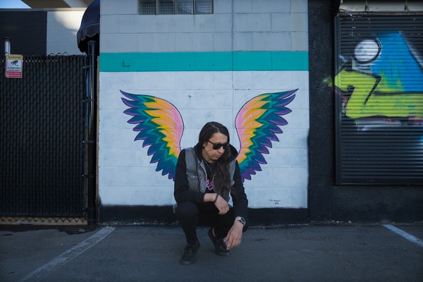 A colour photo shows the artist Sunflower Dreams crouching in front of a wall with wings painted on it, so it looks like they have wings. 