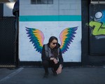 A colour photo shows the artist Sunflower Dreams crouching in front of a wall with wings painted on it, so it looks like they have wings. 