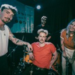 A colour photo shows four members of the band Sunglaciers pose while setting up for a show on stage in front of a black curtain