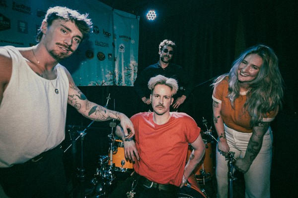 A colour photo shows four members of the band Sunglaciers pose while setting up for a show on stage in front of a black curtain