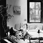 A black and white photo shows artist sunshower laying on his back on a couch in a living room playing an electric guitar