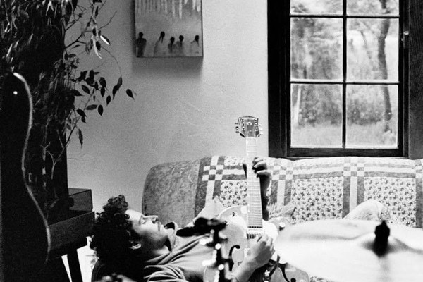 A black and white photo shows artist sunshower laying on his back on a couch in a living room playing an electric guitar