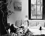A black and white photo shows artist sunshower laying on his back on a couch in a living room playing an electric guitar