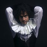 A colour photo shows artist T Gowdy against a black background wearing a ruffled collar and white pearls. They're arms are behind their head. 
