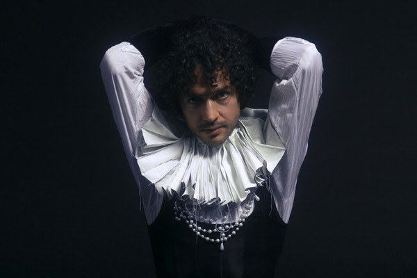 A colour photo shows artist T Gowdy against a black background wearing a ruffled collar and white pearls. They're arms are behind their head. 