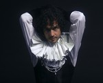 A colour photo shows artist T Gowdy against a black background wearing a ruffled collar and white pearls. They're arms are behind their head. 