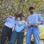 A colour photo shows three-piece band Tawni Bias posing in front of a leafy tree on a sunny day